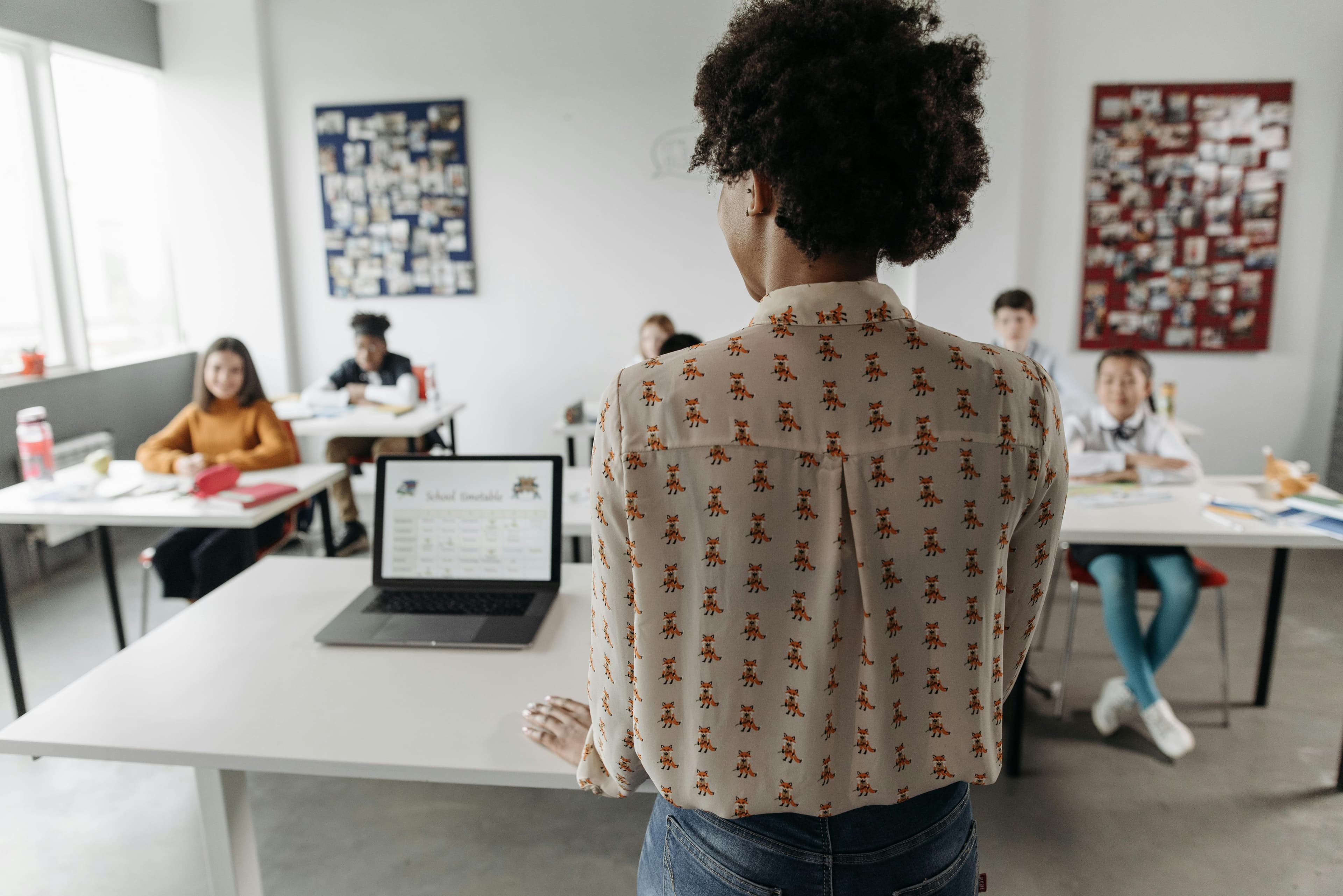 A teacher standing in a classroom with students and a laptop.